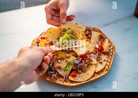 Die Menschen greifen gelbe Mais-Nacho-Chips mit gemahlenem Rindfleisch, Guacamol, geschmolzenem Käse, Paprika und Cilantro-Blättern in Platte auf weißem Stein Stockfoto