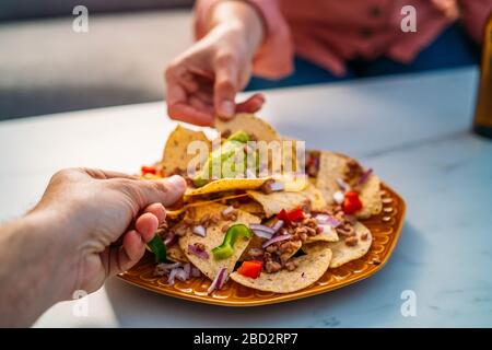 Die Menschen greifen gelbe Mais-Nacho-Chips mit gemahlenem Rindfleisch, Guacamol, geschmolzenem Käse, Paprika und Cilantro-Blättern in Platte auf weißem Stein Stockfoto