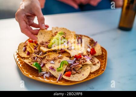 Die Menschen greifen gelbe Mais-Nacho-Chips mit gemahlenem Rindfleisch, Guacamol, geschmolzenem Käse, Paprika und Cilantro-Blättern in Platte auf weißem Stein Stockfoto