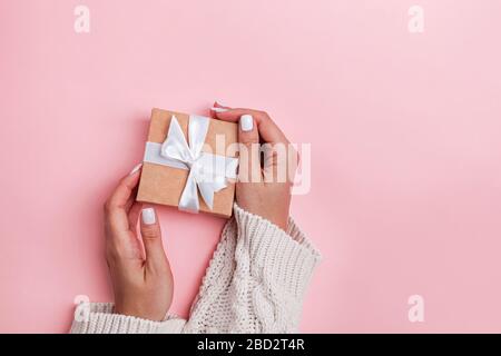 Frauen in der Hand mit Blick auf den oberen Rand, die braune Geschenkbox mit weißem Band auf pinkfarbenem Pastell-Hintergrund halten Stockfoto
