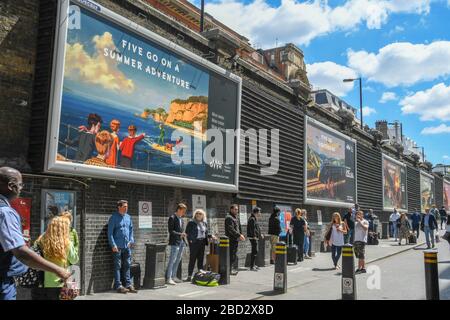 LONDON, ENGLAND - JULI 2018: Werbeplakate an einer Wand außerhalb des Bahnhofs London Paddington mit einer Menschenmenge, die auf dem Weg steht Stockfoto