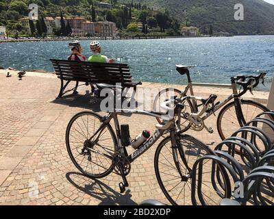 Torbole, Gardasee, Italien - Mai 2017: Zwei Radfahrer machen eine Pause auf einer Seeuferbank, nachdem sie ihre Fahrräder in den Regalen an der Promenade gesichert haben Stockfoto