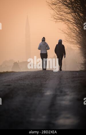 Nahaufnahme von zwei Mädchen, die im frühen Morgenlicht eine unbefestigte Straße entlang gehen, mit einem entfernten Dorf, das von Dunst bedeckt ist Stockfoto