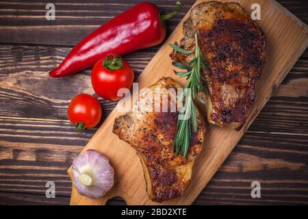 Gegrilltes Schweinesteaks und Gemüse auf einem rustikalen Holztisch. Gegrilltes Fleisch am Knochen mit Gewürzen, Knoblauch, Paprika, Tomaten und Rosmarin. Restaurant-Menü. Ansicht von oben Stockfoto