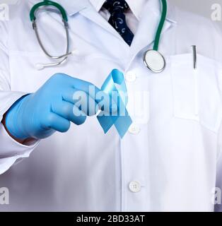 Doktor in einem weißen Mantel und Krawatte steht und hält ein blaues Seidenband in einer Schleife, trägt blaue sterile medizinische Handschuhe, Symbol des Kampfes und der Behandlung o Stockfoto