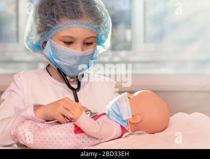 Ein kleines Mädchen sitzt zu Hause während der Quarantäne und spielt einen Arzt, behandelt und hört die Puppe mit einem Stethoskop in einer medizinischen Maske, Hut Stockfoto