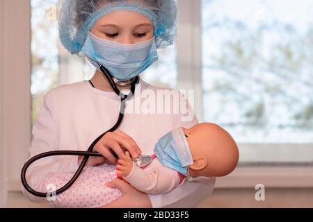 Ein kleines Mädchen sitzt zu Hause während der Quarantäne und spielt einen Arzt, behandelt und hört die Puppe mit einem Stethoskop in einer medizinischen Maske, Hut Stockfoto