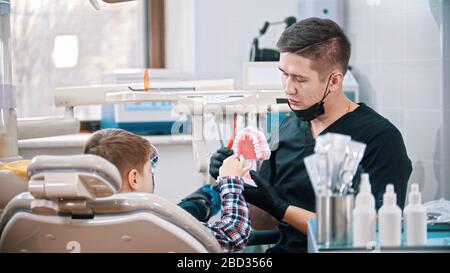 Man-Zahnarzt, der einem kleinen Jungen bei seiner Behandlung ein plastisches Modell der Kiefer zeigt. Mittelaufnahme Stockfoto