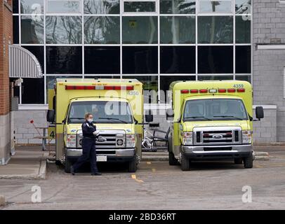 COVID-19 zwei gelbe Ambulanzen, die im Noteingang des Krankenhauses geparkt sind, mit einer Sanitäterin, die eine Maske trägt Stockfoto