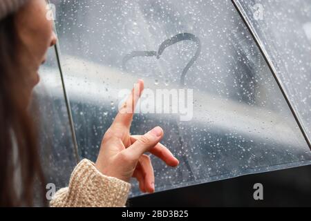 Mädchen malt das Herz auf einer nebligen Oberfläche im Regen unter einem Regenschirm. Love Story Konzept Stockfoto