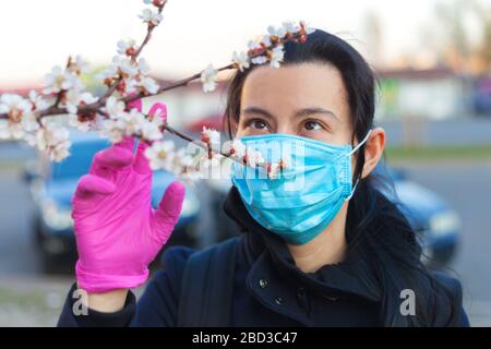 Ein Mädchen in einer schützenden medizinischen Maske und Gummihandschuhen, im Frühling auf der Straße, schaut auf einen blühenden Apfel oder Kirsche. Nahaufnahme Stockfoto