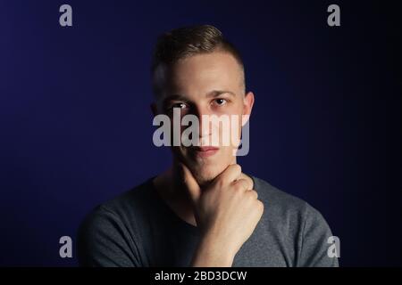 Junger kaukasischer blonder Mann in grauem T-Shirt auf dunkelviolettem Hintergrund, der sein Kinn berührt und an etwas denkt Stockfoto