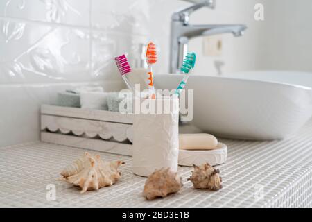 Drei verschiedenfarbige Zahnbürsten stehen in einem Keramikglas vor dem Hintergrund des Waschbassins und anderen Accessoires im Bad. Stockfoto