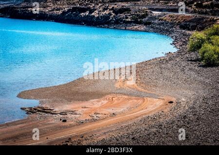 Türkisfarbener See und Strand aus rötlichen Steinen an der Seite einer Straße in Th Stockfoto