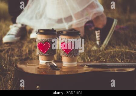 Ein romantisches Paar umarmt, während es bei Sonnenuntergang Picknick machen kann. Kaffeetasse kuscheliger Strickärmel mit filzrotem Herz. Stockfoto