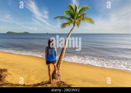 Blick auf einen einzigen weiblichen Tourist, der in der Nähe einer Kokospalme am Clifton Beach mit Blick auf den Pazifischen Ozean und die Doppelinsel in Queensland steht. Stockfoto