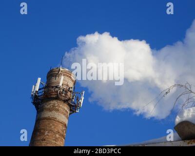 Rohre einer alten Fabrik werfen Wolken von giftigem weißem Rauch in den Himmel, die die Atmosphäre schädigen. Urbaner Smog aus Rauch aus Kesselhäusern. Weißer Rauch aus einem Kamin gegen einen blauen klaren Himmel. Stockfoto