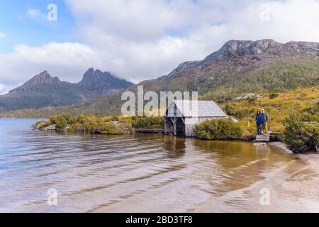 Blick von der Küstenlinie zweier Wandertouristen am Dove See von Cradle Mountain und seinem berühmten Bootshaus. Stockfoto