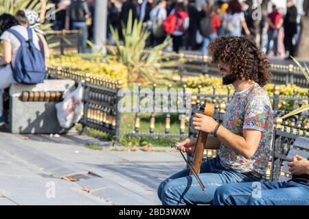 Der junge Mann mit geschweiften Haaren auf der Straße singt mit einem Geigeninstrument. Er sitzt auf der Bank. Die Leute geben Geld. Stockfoto