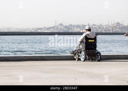 Ein behinderter Mann sitzt im Rollstuhl am Strand. Er beobachtet das Meer. Die Böden bestehen aus Beton und werden an einem sonnigen Tag ins Freie gebracht. Stockfoto