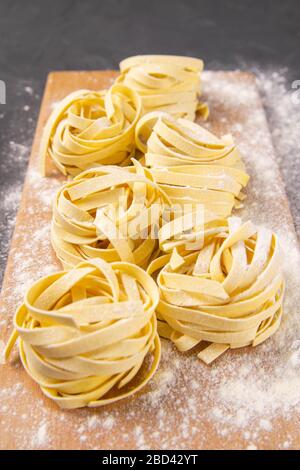 Verschiedene italienische hausgemachte rohe, ungekochte Pasta Spaghetti und Tagliatelle mit Mehl auf Holzbrett über schwarzem Texturhintergrund. Flaches Lay Stockfoto