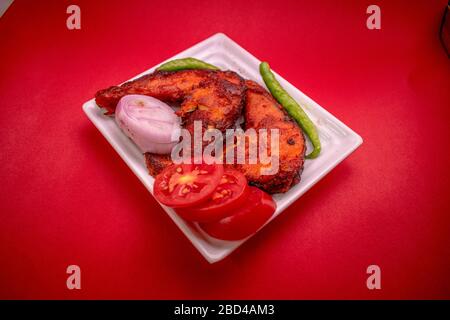 Gebratener Fisch in weißem Teller mit grünem Chili, Zwiebeln und Tomaten auf rotem Hintergrund Stockfoto