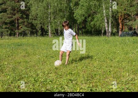 9-jähriger Junge in Uniform spielt im sonnigen Sommertag Fußball auf grüner Wiese, Kind tritt Fußball, wirft es Stockfoto