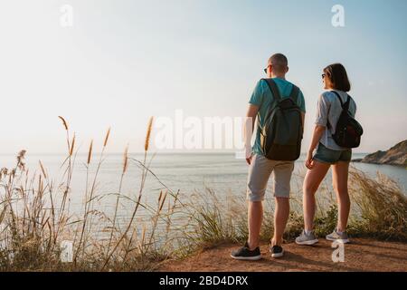 Ein paar junge Wanderer mit Rucksäcken stehen am Meeresblick und genießen den Sonnenuntergang Stockfoto