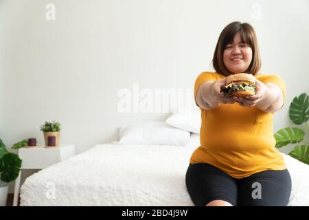 Hungrig übergewichtige Frau und lächelnd Hamburger und Sitzen im Schlafzimmer, sie sehr glücklich und genießen Sie Fast Food zu essen. Konzept der Essattacken d Stockfoto