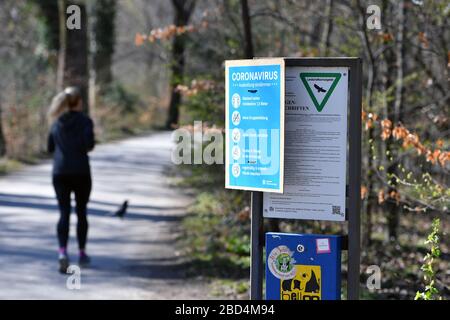 Das öffentliche Leben in Zeiten der Coronavirus-Pandemie am 5. April 2020 im Englischen Garten in München. Am Eingang zum Park hängt ein SCHILD MIT Verhaltensregeln VON CORONA. Dahinter läuft ein Freizeitsportler Jogger. Das frühlingshafte Wetter treibt die Bevölkerung in die frische Luft, immer mit der notwendigen Entfernung aufgrund einer Infektion mit dem Corona-Virus. Ausstiegsbeschränkungen, Kontaktsperre. Weltweite Verwendung Stockfoto