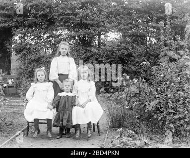 Ein klassisches frühes, englisches Schwarz-Weiß-Foto von Edwardian, das vier Schwestern zeigt, die für den Fotografen in ihrem hinteren Garten posieren. Die älteste Schwester, die hinten steht, die beiden mittleren Schwestern sitzen auf Stühlen, die jüngste Schwester steht dazwischen. Stockfoto