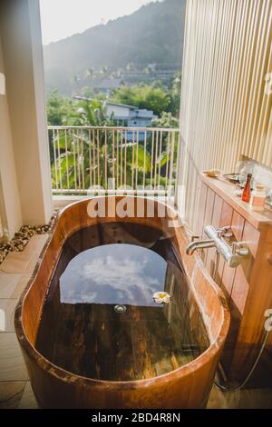 Braune Holzbadewanne voll mit Wasser auf dem Balkon mit malerischer Aussicht auf grüne Dschungel Palmen in Ao Nang Dorf in Krabi Provinz, Thailand Stockfoto