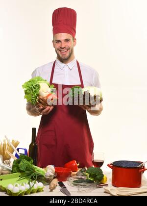 Konzept des Kochvorgangs. Mann mit Bart isoliert auf weißem Hintergrund. Kochen Sie mit geschirr und Zutaten am Tisch. Der Koch in Burgunderuniform hält Körbe mit Kohl und Salat. Stockfoto