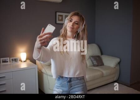 Lustige Mädchen mit Telefon und großen Kopfhörern machen ein Foto auf ihrem Smartphone. Eine junge schöne Frau in Jeans und ein weißer Pullover nimmt ein selfie. Stockfoto