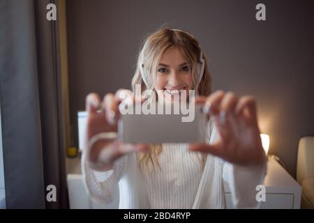 Lustige Mädchen mit Telefon und großen Kopfhörern machen ein Foto auf ihrem Smartphone. Eine junge schöne Frau in Jeans und ein weißer Pullover nimmt ein selfie. Stockfoto