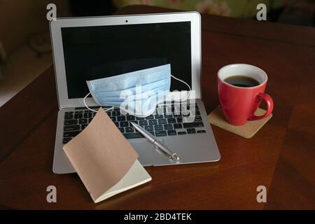 Arbeiten Sie von zu Hause aus während einer Kovid-19-Pandemie. Konzentrieren Sie sich auf die chirurgische Gesichtsmaske auf dem Laptop und eine rote Tasse Kaffee auf dem Esstisch. Draufsicht. Stockfoto