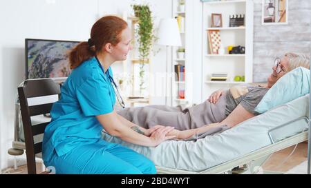 Weibliche Assistentin aus dem Pflegeheim, die die Hand der alten Frau im Krankenbett liegt und mit ihr spricht Stockfoto