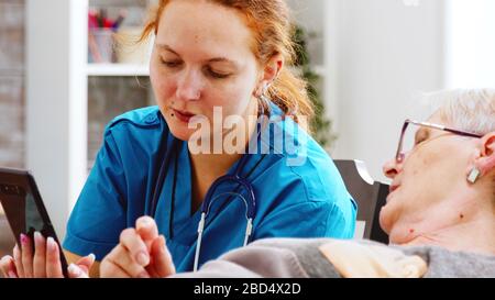 Nahaufnahme der Krankenschwester, die einer älteren behinderten Frau hilft, ein Smartphone zu benutzen. Helles Zimmer mit großen Fenstern Stockfoto