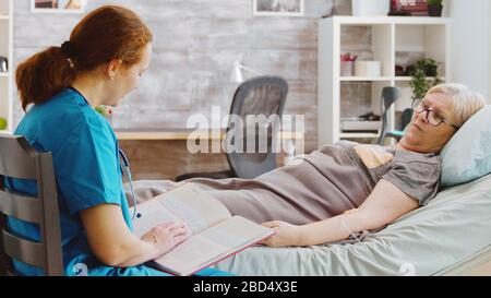 Aufschlussreiche Aufnahme einer alten Dame, die im Krankenbett liegt, während eine Krankenschwester ihr ein Buch vorliest. Betreuer und Sozialarbeiter Stockfoto