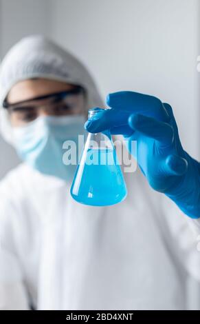 Forscher tragen blaue Latexhandschuhe und Coronavirus Schutzkleidung Blick in einen Kolben der blauen Flüssigkeit Stockfoto