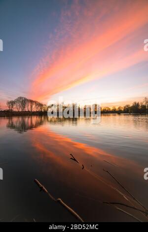 Wolken werden durch das Licht der untergehenden Sonne bunt beleuchtet. Schöne Reflexionen im ruhigen Wasser des Sees. Stockfoto