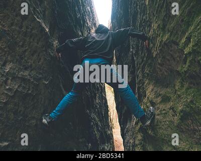 Lustiger Mann, der zwischen Felswand klettert. Mann trägt blaue Hose und schwarze Jacke. Stockfoto