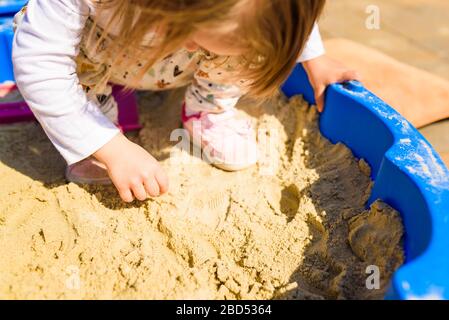 Kleines Mädchen, das an einem sonnigen Tag im Freien in einer Sandbox spielt. Kind im Sommer in blauer Sandbox. Stockfoto
