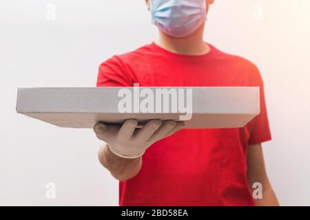Der Lieferer trägt ein rotes T-Shirt, Schutzhandschuhe und eine Maske, die den Pizzakasten in der Nähe hält. Stockfoto