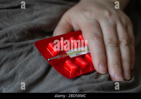 Blister mit Pillen in weiblicher Hand. Weibliche Handfläche ruht auf einer Blisterpackung mit Pillen. Das Thermometer befindet sich in der Nähe. Draufsicht in einem Winkel. Selektiver Fokus. Stockfoto