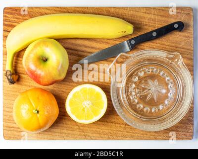 Stillleben von Fruchtsalatzutaten apfelorange und Banane mit einer halben Zitrone und einem Küchenmesser und Zitronenquetschen auf einem Holzhackbrett Stockfoto
