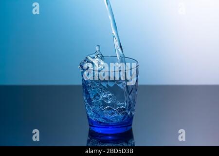 Sauberes Wasser wird in ein Glas gegossen, das auf einem schwarzen Tisch steht. Blaues Glas mit fließendem Wasser, das auf einem schwarzen Tisch auf blauem Grund steht Stockfoto