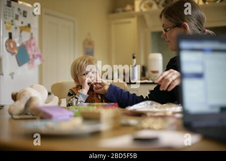 Mutter füttert ihre Kinder beim Essen am Küchentisch während der Arbeit von zu Hause während der Zeit der Selbstisolation - 2020 COVID-19 Coronavirus Pandemie Stockfoto