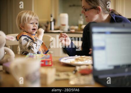 Mutter füttert ihre Kinder beim Essen am Küchentisch während der Arbeit von zu Hause während der Zeit der Selbstisolation - 2020 COVID-19 Coronavirus Pandemie Stockfoto