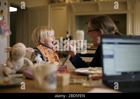 Mutter füttert ihre Kinder beim Essen am Küchentisch während der Arbeit von zu Hause während der Zeit der Selbstisolation - 2020 COVID-19 Coronavirus Pandemie Stockfoto